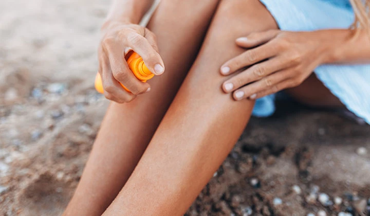 Frau am Strand sprüht Sonnenspray auf ihre Beine 